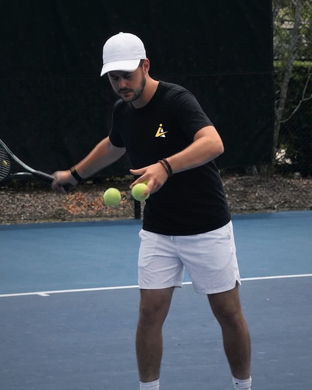 Alex Navrotchi feeding tennis balls on a blue hard court, racquet in hand, Inner Ace T-shirt